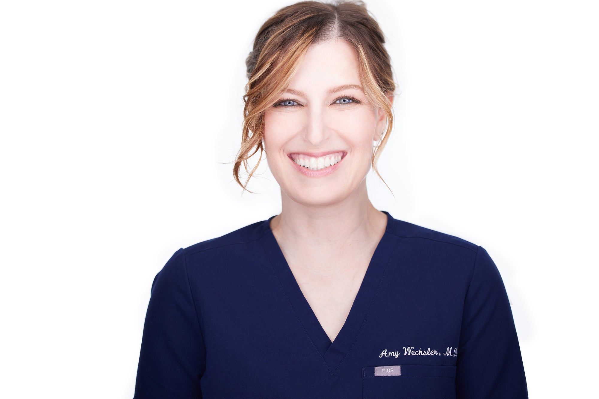 Dr. Amy Wechsler wears navy blue scrubs. Her hair is tied back and a two wispy bang fall down on either side of her face. She smiles.