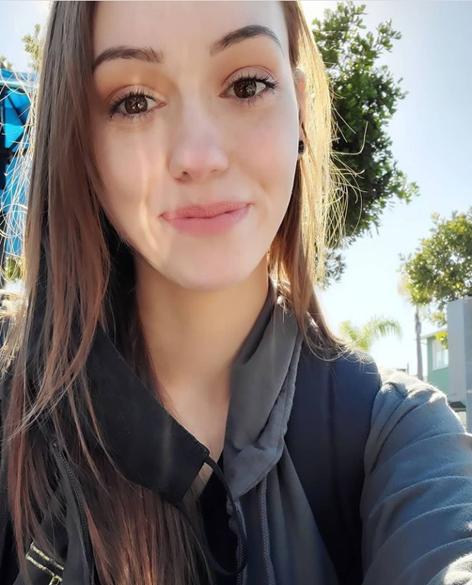 Jessica smiles gratefully at as as she takes a selfie. Trees are behind her, and the sun shines down on her.
