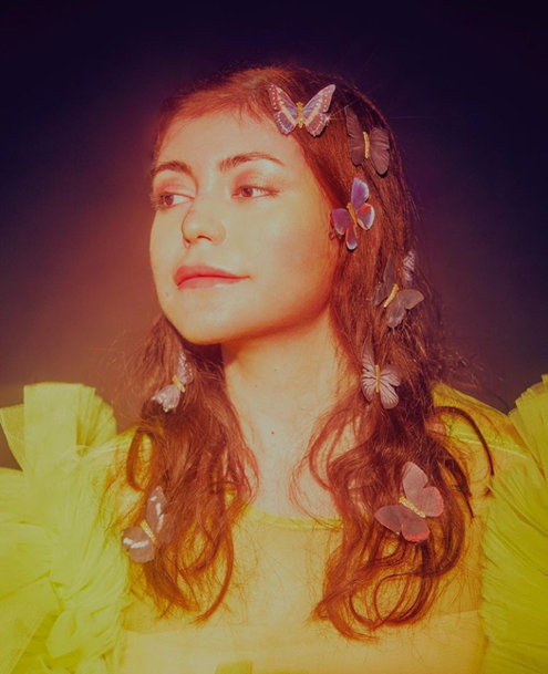 A radiant and angelic light is cast upon Ariana, butterflies flutter on her hair, and she is wearing a yellow dress. She smiles calmly.