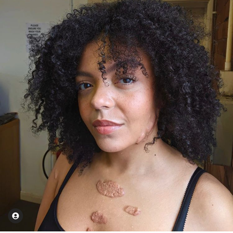 Bianca has free flowing curly hair that pushes outwards and then down. Here, she wears a black tank top, and gives a small and candid smile like she is in the middle of a thought.