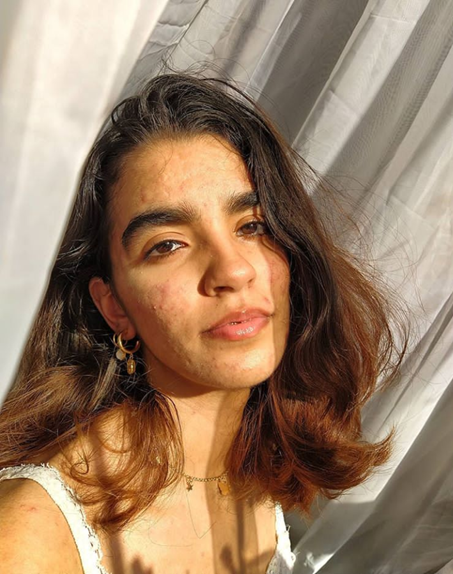 Constanza sits upright with relaxed shoulders, and her head is tilted back slightly. Her wavy, brown, and black hair falls playfully at her shoulders. The sun shines on her.