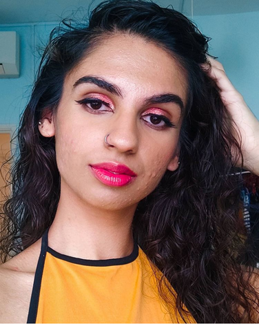 Davina is wearing electric pink lipstick, electric pink eyeshadow, and she has wavy, dark brown, and long hair. She wears an orange halter top.