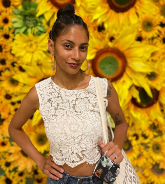 Destiny is standing in front of a back drop of sunflowers. She has brown skin with red undertones and deeply colored black hair. She is wearing a sleeveless white lace crop top, hoops, maroon lip stick, and a top bun. 