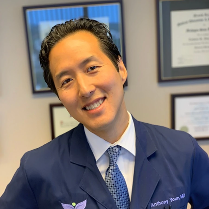 Dr. Anthony Youn wears a navy blue doctor's coat, a white button down shirt, and a tie. He places his hands on his hips, tilts his head to the left kindly, and he smiles cordially. His credentials are on the wall behind him.