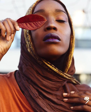Fanta wears deeply colored brown hijab with gold trimming, an orange tunic, and plum lipstick. She holds a brown leaf over her left eye. Her eyes are closed as she lifts her chin.