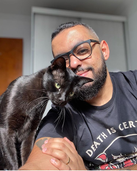 Ibrahim wears sunglasses with black rims, and has neatly cut hair. He smiles as he hold his face next to his black cat. The cat leans into him lovingly. The sun is shining.
