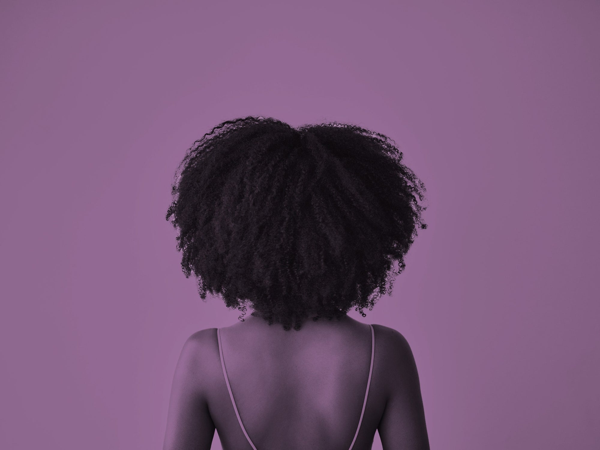 An African-American girl with voluminous, thick hair that hangs stands with her back towards us. The photo has a purple hue.