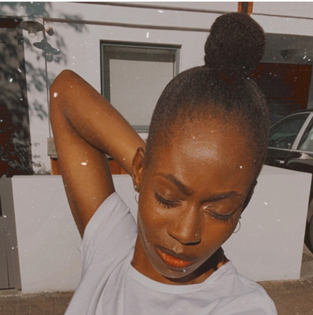 Joanne has deeply colored brown skin, and she glances down with her hand behind her head. She appears to be focusing or finding her center.