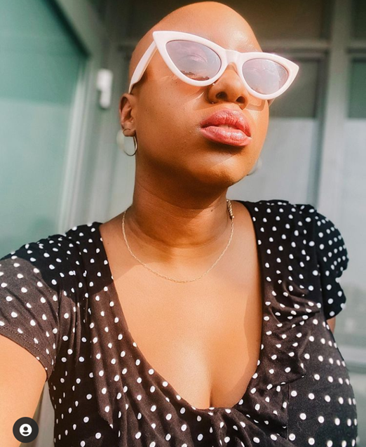 Joy is facing the sun and wearing white pointed tip sun glasses. She is wearing a black dress with white polka dots and hoops. She looks undaunted and confident. 