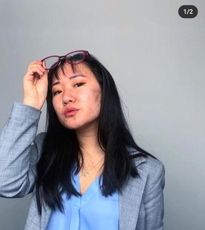 Katie is wearing a blue V-neck blouse, a grey blazer, and she has thick, black hair that is falling past her shoulder. She lift her red glasses upwards as if she is about to put them in her hair. She looks out observantly.
