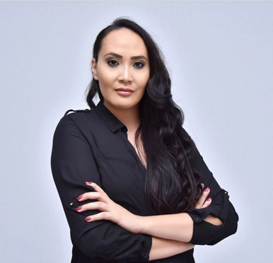 Kelly Holmes has deeply tan skin, prominent cheek bones, and powerful eyes. She has dark brown and very long hair. She is crossing her arms and is looking at us with strength, determination, and confidence. She is wearing bold, red fingernail polish.