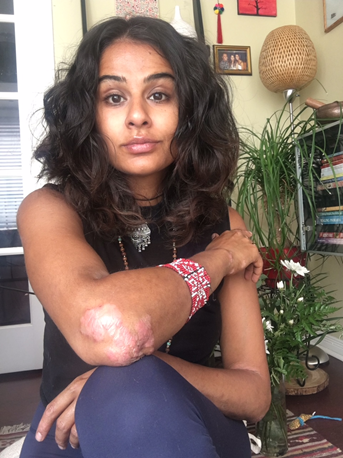 Reena has brown skin, and she is looking at us as candidly with her eyebrows raised. The left corner of her mouth is raised like she is about to smile. She has brown eyes, and dark brown hair with thick waves falling just past her shoulders.