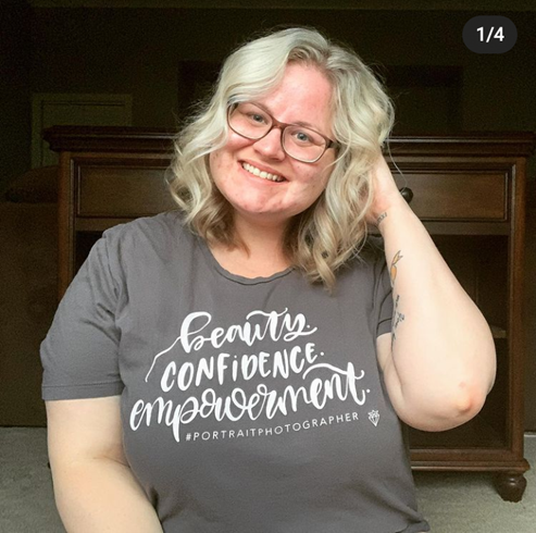 In this picture, Sarah has curly, whitish, and blonde hair that is cut into a bob. She wears a T-shirt that reads: beauty, confidence, and empowerment. Her touches her right hand to her hair joyfully.