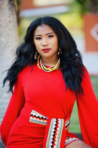 The picture shows Dr. Thomasina standing powerfully. She wears a bright red dress, red lipstick, a beaded necklace, and the the dress that ties in the front has indigenous print.