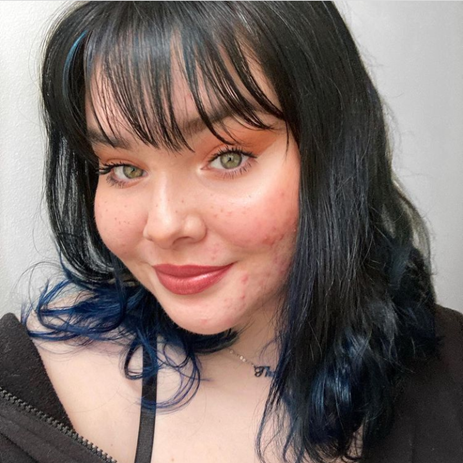 Theresa has black hair with blue ends. She has prominent cheek bones and green eyes. She raises her eyebrows, and the right side of her mouth raises into a smile. 