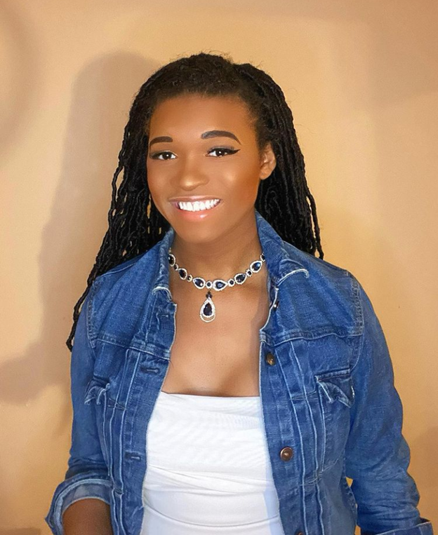 Xay has long and wavy braids. She wears a grey tank top, a jean jacket, and a silver and blue necklace. She smiles with a happy friendliness.
