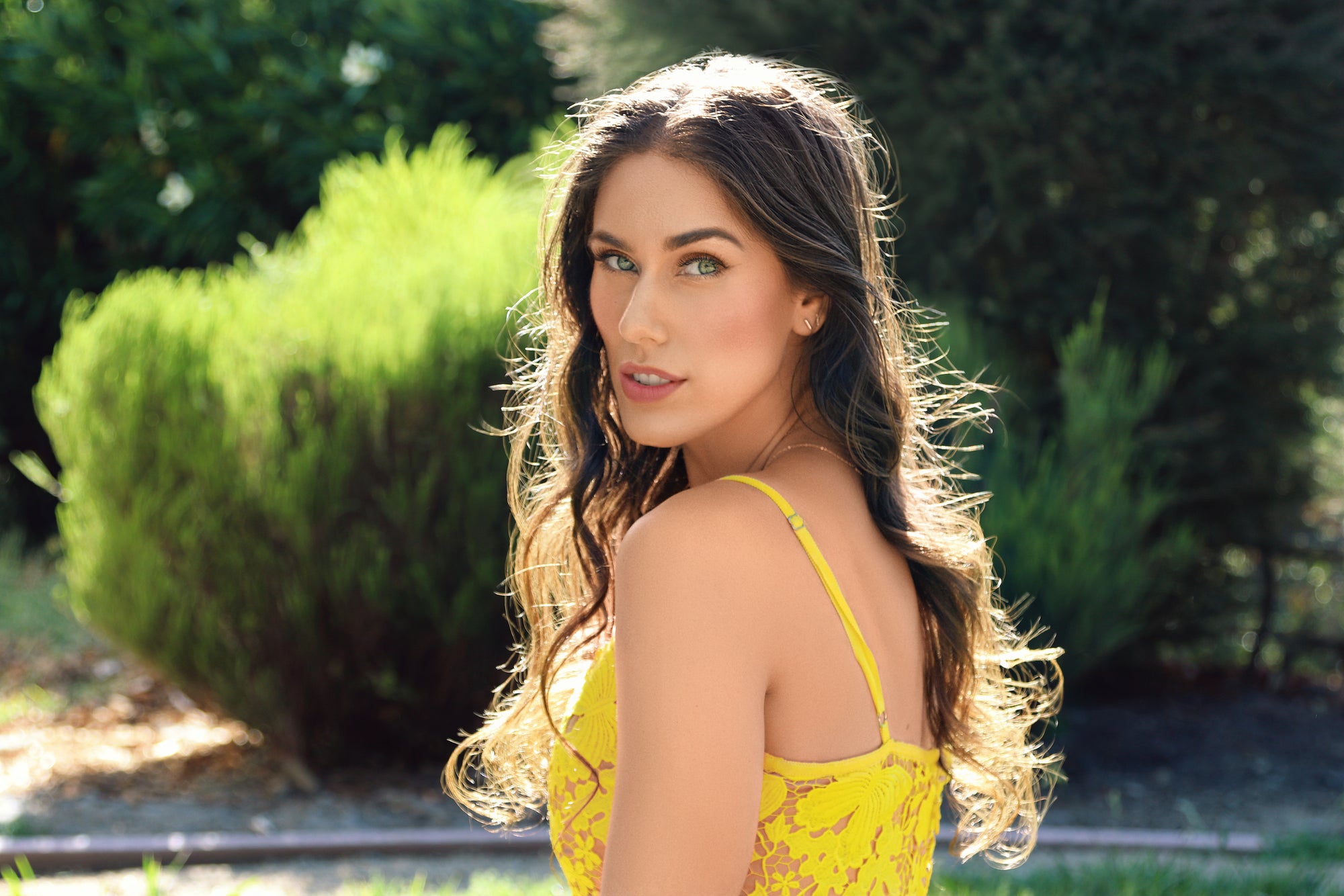 Cassandra bankson sitting on the grass at a park with sunlight falling through hair in a yellow lace dress with butterflies looking over shoulder wearing spf makeup